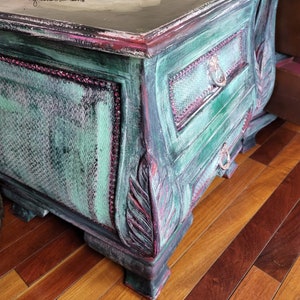 May include: A distressed green and black painted bedside table with ornate carvings and a drawer. The drawer has a metal pull and a decorative metal ring. The bedside table is sitting on a wooden floor.