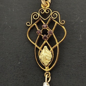 May include: A gold-toned pendant necklace featuring an openwork metal frame with scrollwork at the top, three small purple stones set in a diamond pattern, a central textured leaf motif, and a single white baroque pearl drop at the bottom.
