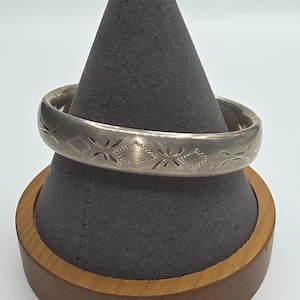 May include: A silver-toned hinged bangle bracelet featuring a repeating engraved pattern of diamond shapes and stylized cross motifs. The metal surface has a matte finish with visible signs of wear, and the design wraps around the entire band.