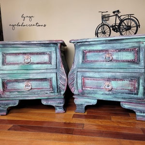 Op de afbeelding: Een paar nachtkastjes in distressed groene kleur met elk twee laden. De nachtkastjes hebben sierlijke houtsnijwerk en decoratieve metalen beslag. De tekst "by aga agalobocreations" is zichtbaar op de muur achter de nachtkastjes.