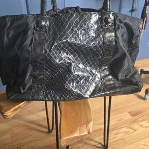 May include: A black tote bag featuring a central panel with a glossy, textured snakeskin pattern. The sides of the bag are made of a smooth, matte black fabric. It includes two matching black shoulder straps and a button closure at the top.