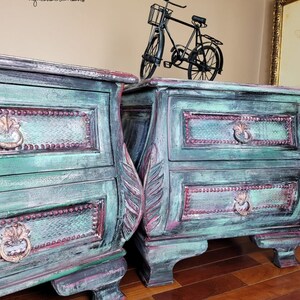 May include: Two antique-style bedside tables with two drawers each. The bedside tables are painted in a distressed green and have ornate carvings. The bedside tables are sitting on a wooden floor.