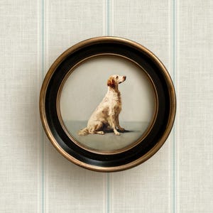 May include: A round framed dog portrait. The artwork features a seated dog with a light brown and white coat, set against a soft gray background. The frame is black with a gold-colored inner rim. The artwork is displayed against a striped wallpaper.