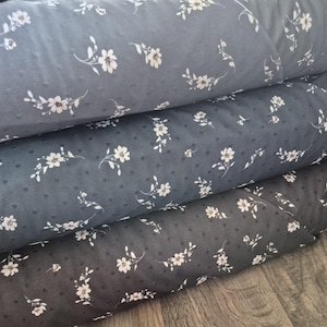 May include: Close-up of several rolls of fabric in varying shades of gray. Each roll is adorned with a delicate white floral pattern and small, evenly spaced dots. The fabric appears soft and textured, suitable for various sewing projects.