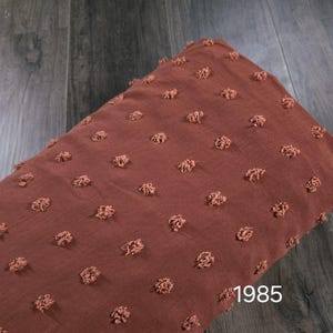 May include: A folded piece of brown fabric with a textured, dotted pattern. The fabric has a soft, slightly wrinkled appearance. The number "1985" is visible in the lower right corner. The fabric is likely used for clothing or home decor.