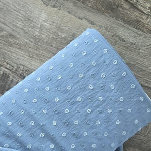May include: A folded piece of light blue fabric with a textured, dotted pattern. The fabric has a soft appearance and is likely used for clothing or home decor projects. The dots are a lighter shade of blue.