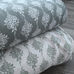 May include: Two rolls of fabric with a floral pattern. The top roll is a muted gray with white rose bouquets and vines. The bottom roll is white with a similar gray floral design. The fabric is folded and stacked.