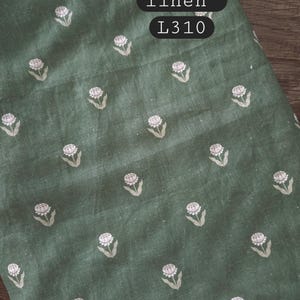 May include: A piece of sage green fabric featuring a repeating pattern of small white flowers. The text 100% stonewashed linen L310 is printed in black boxes on the fabric. The material has a visible woven texture throughout the surface.