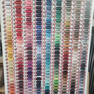 May include: A wall display of spools of thread in various colors, organized by color family. The spools are arranged in rows and columns, with each row containing a different color. The colors include red, orange, yellow, green, blue, purple, pink, brown, and white.