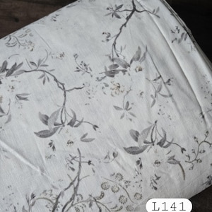 May include: A folded bolt of off-white fabric featuring a printed pattern of gray botanical branches, leaves, and small flowers. A white rectangular label with the black text L141 is attached to the lower right corner of the textile.