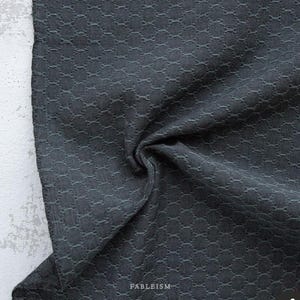 May include: Dark gray fabric with a textured, repeating pattern. The fabric is draped, showing the texture and depth of the material. The word "FABLEISM" is printed in white at the bottom right corner.