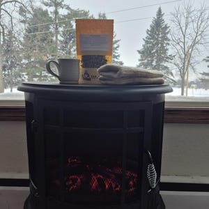 May include: A black electric fireplace with a mesh front, topped with a mug, a package of tea, and a pair of mittens. The tea package has text on it. The background shows a snowy landscape.