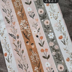 May include: A fabric featuring vertical stripes with a floral and acorn pattern in shades of orange, white, and muted green. The textile includes the printed text 100% cotton and 2427. The design consists of repeating botanical illustrations of wildflowers and small acorns.