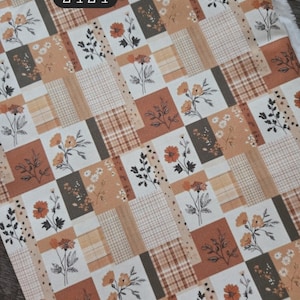 May include: A 100% cotton fabric featuring a patchwork pattern of rectangular blocks in shades of cream, tan, brown, and orange. The design includes floral illustrations, sprigs of leaves, and plaid grid patterns. Text reads 100% cotton 2424.