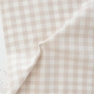 May include: A close-up of a beige and white gingham fabric. The fabric is folded, showing the texture and pattern. The fabric is a light, neutral color, suitable for various sewing and crafting projects. The word "FABLEISM" is printed on the bottom right.
