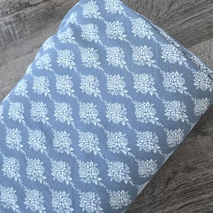 May include: A folded piece of blue fabric with a repeating pattern of white roses and leaves. The floral design is arranged in diagonal rows across the material. The fabric appears soft and slightly textured.