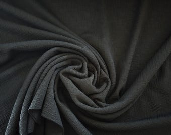 May include: A dark gray fabric with a textured, woven pattern. The fabric is draped and swirled, creating folds and shadows. The material appears to be soft and flowing, suitable for clothing or home decor.
