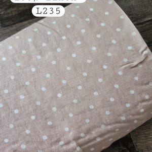 May include: A roll of beige fabric featuring a white polka dot pattern. The fabric is labeled with the text "stonewashed", "100% linen", and "L235". The material has a visible woven texture and is displayed in a rolled format.