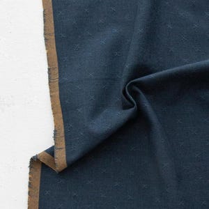 May include: A dark navy blue fabric featuring a subtle, repeating cross-stitch pattern across the surface. The material has a distinct brown selvage edge along the side, and the word FABLEISM is printed in white at the bottom center of the textile.