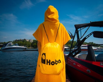 May include: A person wearing a yellow hooded towel with a yellow drawstring bag that says "Hobba" on it. The person is standing on a dock with a red and black boat in the background.