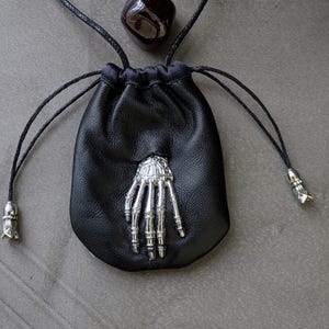 Peut inclure: Pochette noire en cuir avec un emblème de main de squelette argenté. La pochette a un cordon noir et des accents argentés. Une pierre précieuse rouge foncé et une pierre rose sont également visibles.