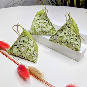 May include: Three green triangular sachets with white lace detailing. The sachets are on a white surface with dried flowers.