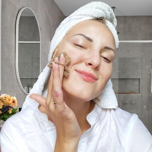 May include: A woman with a white towel wrapped around her head applies a brown facial mask to her cheek.
