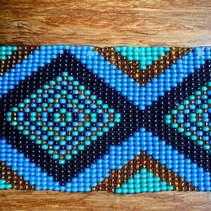May include: Close-up of a beaded bracelet or belt with a geometric pattern. The design features a repeating pattern of blue, teal, black, and gold beads. The beads are tightly woven together, creating a textured surface. The background is a wooden surface.