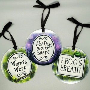 May include: Three round ornaments with black ribbon hangers. Each ornament features a different phrase: "Worm's Wort", "Deadly Night Shade", and "Frog's Breath". The ornaments have a watercolor-style background with green, purple, and white accents.
