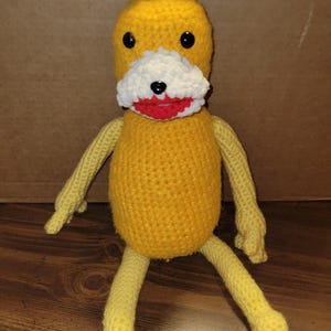May include: A handmade yellow crochet duck toy with black button eyes, a white and red mouth, and yellow arms and legs. The duck has a tuft of yellow hair on top of its head. The toy is sitting on a wooden surface.