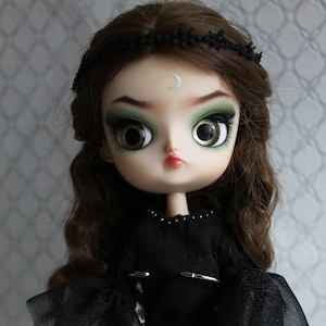 May include: A doll with long brown hair wearing a black dress with a sheer overlay and a silver crescent moon on her forehead.