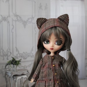 Könnte beinhalten: Eine Puppe mit langen grauen Haaren, die einen braunen und roten karierten Mantel mit einer Katzenohrenhaube trägt. Die Puppe hat grüne Augen und trägt unter dem Mantel ein weißes Kleid.