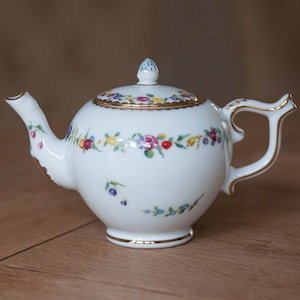Könnte beinhalten: Eine weiße Porzellan-Teekanne mit floralem Muster und goldenen Akzenten. Die Teekanne hat einen runden Korpus und einen geschwungenen Ausguss. Der Deckel hat einen kleinen Knopf oben.