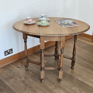 May include: A round wooden drop-leaf table with a medium brown finish and turned legs. On the tabletop sit three porcelain teacups with matching saucers in pink, blue, and green, along with a magazine titled Decanter.