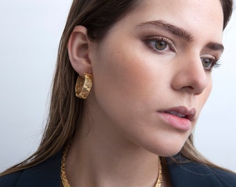 May include: A woman wearing gold hoop earrings with a textured design. The earrings are large and have a circular shape.