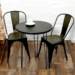 May include: A black bistro set with a round table and two matching chairs. The table has a dark wood top and hairpin legs. A white teacup and saucer sit on the table. The chairs have a metal frame and a slatted back.