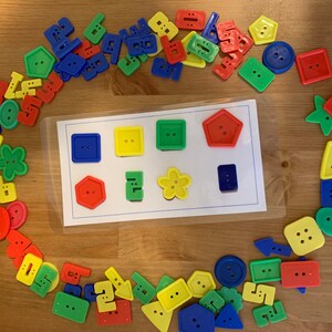 May include: A set of colorful plastic buttons in various shapes, including squares, circles, stars, and flowers. The buttons are arranged in a circle around a white card with a grid of nine empty spaces. The card is labeled with the words "Shape Match".