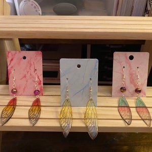 Op de afbeelding: Drie paar oorbellen met kleurrijke, doorschijnende vleugels. De vleugels zijn gelaagd in verschillende kleuren, waaronder geel, groen en rood. De oorbellen worden gepresenteerd op roze en blauwe kaart.