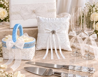 Könnte beinhalten: Ein Hochzeits-Set bestehend aus einem weißen Gästebuch mit Perlen-Schleife, einem weißen Ringkissen mit Spitze und Bändern, einem blau-weißen Blumenmädchen-Korb, zwei Champagnergläsern mit Schleifen sowie einem Tortenmesser und Tortenheber.