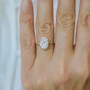 May include: A gold ring with a large, oval-cut diamond. The diamond is set in a four-prong setting and is the focal point of the ring. The band is a simple, polished gold. The ring is on a hand.