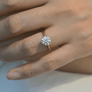 May include: A gold ring with a large, round, clear gemstone set on a delicate band. The ring is on a person's finger, with the hand in soft focus. The ring is a classic design, likely for an engagement or special occasion.