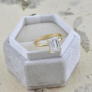 May include: A gold ring featuring a rectangular emerald-cut clear gemstone set in a four-prong setting, resting inside a light gray hexagonal velvet ring box with a white ribbed interior lining and a textured fabric exterior finish.