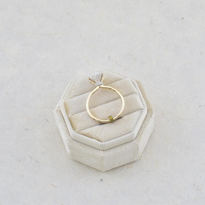 Puede incluir: Un anillo de oro con una gema transparente de talla brillante redonda en un engaste de garras. La banda es fina y de oro pulido, y el engaste incluye un peque&ntilde;o grupo de diminutas piedras de acento visibles bajo la piedra central.