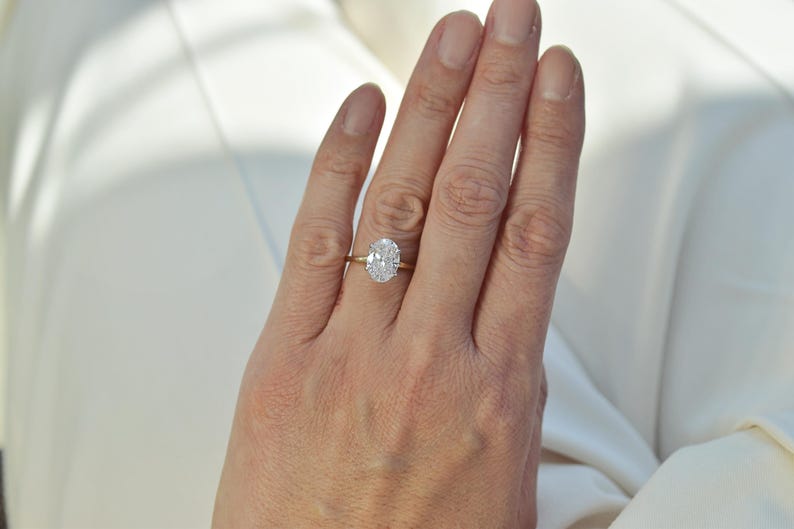 Puede incluir: Un anillo de oro con una piedra preciosa transparente de corte ovalado en un engaste de cuatro garras. La banda es fina y pulida, con un acabado amarillo met&aacute;lico. La piedra preciosa est&aacute; situada en el centro y refleja la luz con facetas.