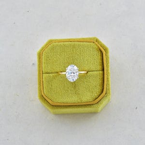 Puede incluir: Un anillo de oro con una gema grande, transparente y de corte ovalado en un engaste de garras. El anillo est&aacute; centrado dentro de un joyero cuadrado de terciopelo color verde chartreuse que tiene una tapa abatible y una ranura horizontal.