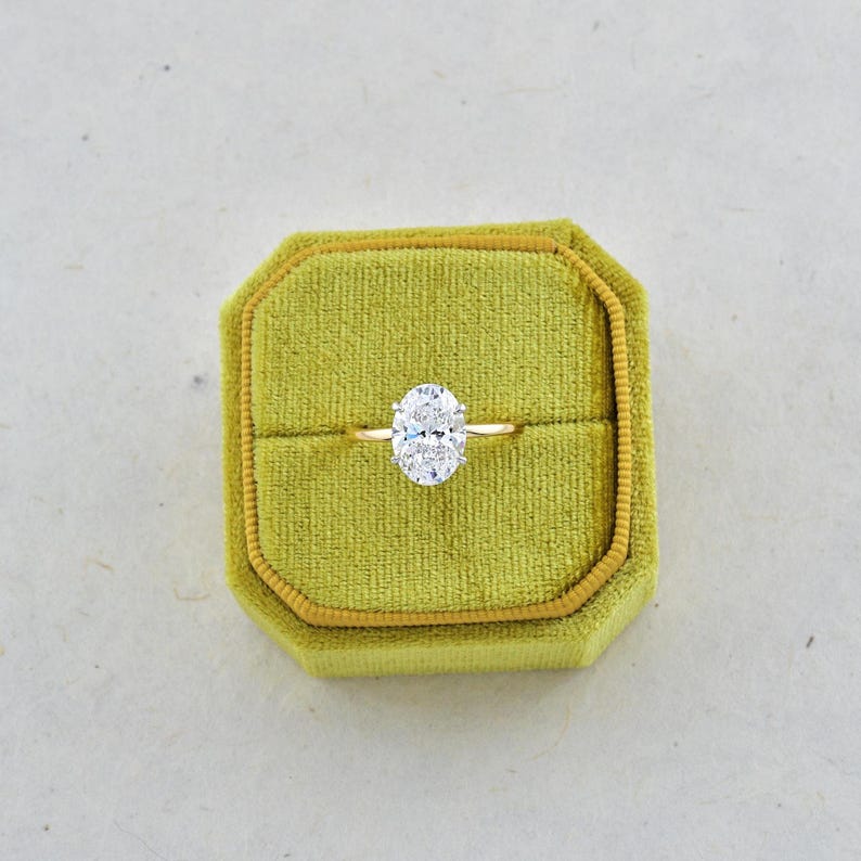 Puede incluir: Un anillo de oro con una gema grande, transparente y de corte ovalado en un engaste de garras. El anillo est&aacute; centrado dentro de un joyero cuadrado de terciopelo color verde chartreuse que tiene una tapa abatible y una ranura horizontal.