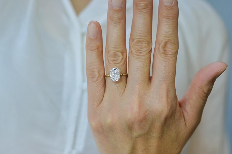 May include: A gold ring with a large, oval-cut diamond. The diamond is set in a four-prong setting and is the focal point of the ring. The band is a simple, polished gold. The ring is on a hand.