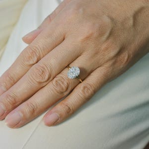Puede incluir: Un anillo de oro con una piedra central grande de forma ovalada rodeada por un halo de piedras m&aacute;s peque&ntilde;as, redondas y transparentes. La banda es fina y pulida, mostrando un dise&ntilde;o cl&aacute;sico con una pieza central brillante y destacada.