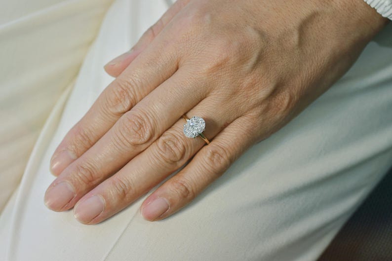 Puede incluir: Un anillo de oro con una piedra central grande de forma ovalada rodeada por un halo de piedras m&aacute;s peque&ntilde;as, redondas y transparentes. La banda es fina y pulida, mostrando un dise&ntilde;o cl&aacute;sico con una pieza central brillante y destacada.