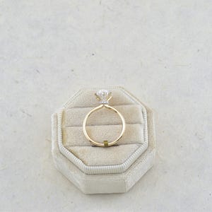 Puede incluir: Un anillo de oro con una piedra preciosa transparente de talla brillante redonda en un engaste de cuatro garras. El aro es fino y de oro pulido. El anillo est&aacute; colocado en posici&oacute;n vertical en un joyero octogonal de terciopelo beige.